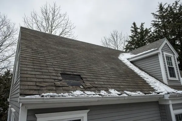 Gray New England house, leftover snow along the eaves and dormer, and a clear gap where shingles tore off, exposing the dark underlayment.